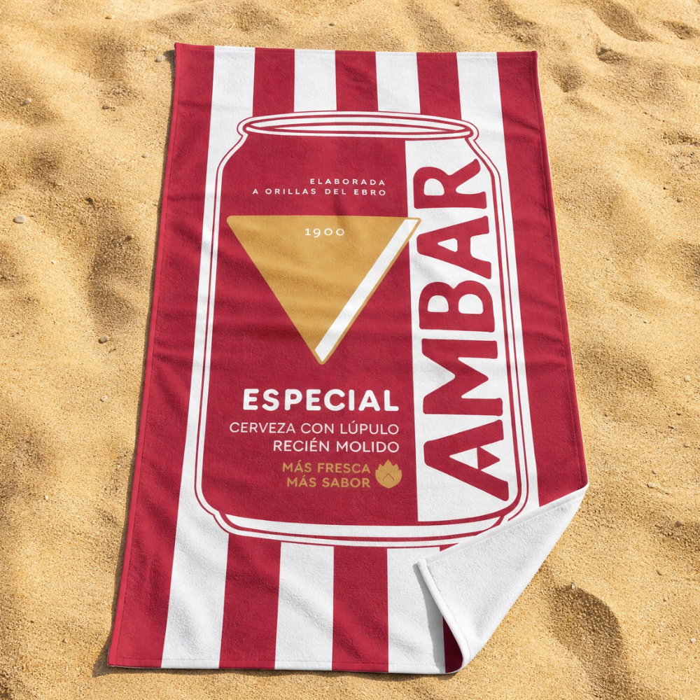 Toalla de microfibra con rayas blancas y rojas y estampado a todo color del diseño de una lata de Cerveza Ambar, extendida sobre la arena de la playa.
